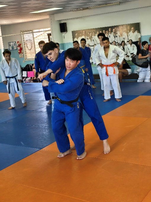 Un judoka con judogi azul y cinturón negro ejecutando la entrada técnica de Seoi Nage sobre un compañero durante una sesión de entrenamiento en el dojo.