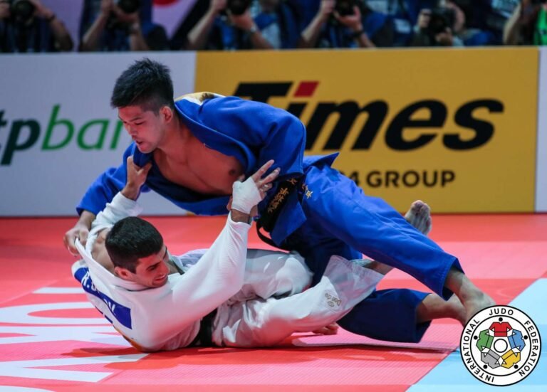 Dos competidores de judo en una transición de técnica de pie a control en el suelo durante un combate.