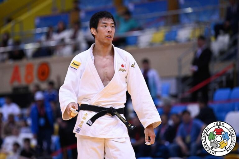 Un plano de tres cuartos del judoka japonés Takanori Nagase de pie sobre el tatami en una arena de judo llena. Viste un judogi blanco y un cinturón negro.