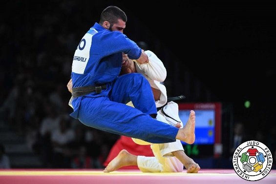El judoka Takanori Nagase con judogi blanco realizando una entrada de proyección baja contra un oponente con judogi azul durante un combate oficial.