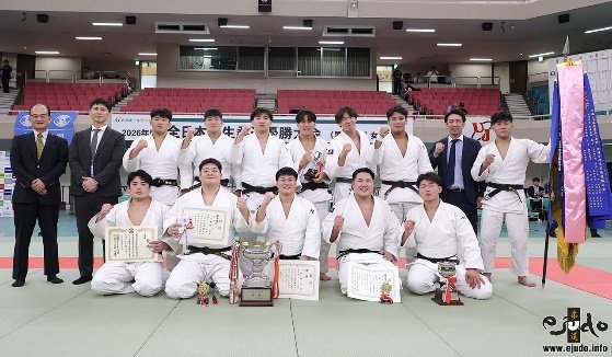 El equipo masculino de judo de la Universidad de Meiji posando con trofeos, diplomas y estandartes tras ganar el campeonato nacional universitario sobre el tatami.