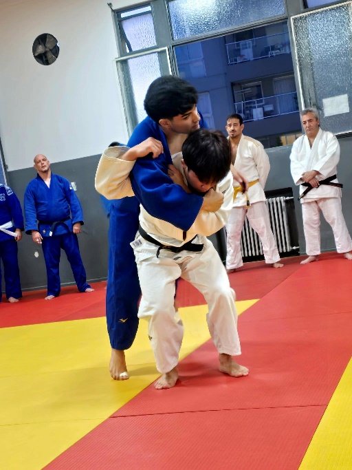 Un judoka con judogi blanco y cinturón negro ejecutando la entrada técnica y desequilibrio de Seoi Nage sobre un compañero con judogi azul durante una práctica en el dojo