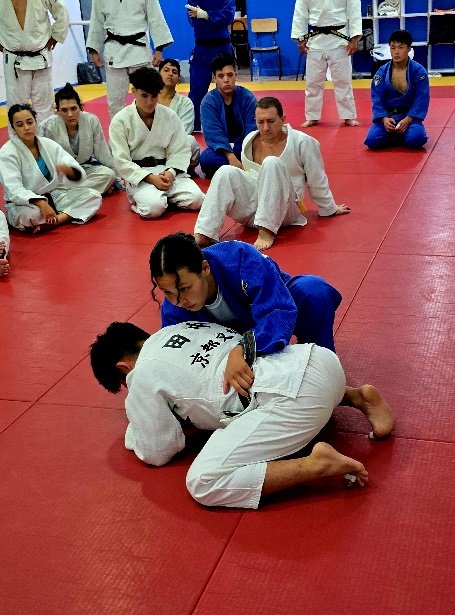 Una judoka con judogi azul ejecutando una técnica de control en suelo sobre un compañero en posición defensiva en el tatami rojo mientras el grupo observa.