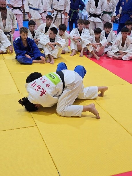 Dos judokas demuestran una técnica de suelo (ne-waza) sobre el tatami mientras un grupo de estudiantes y profesores observa atentamente la explicación técnica.
