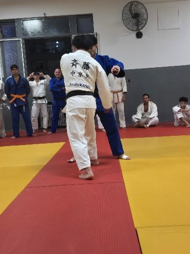 Un judoka con judogi blanco y cinturón negro de espaldas disputando el agarre de Kumi-kata frente a su compañero con judogi azul durante un combate de práctica.