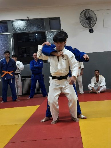 Un judoka con judogi blanco y cinturón negro ejecutando la entrada frontal de Seoi Nage cargando el agarre sobre su compañero con judogi azul durante un entrenamiento en el dojo.