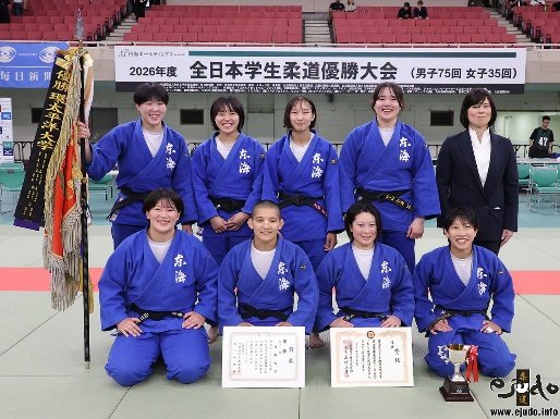 El equipo femenino de judo de la Universidad de Educación de Osaka posando con judogi blanco, trofeos, diplomas y un estandarte junto a su entrenador en el Nippon Budokan.