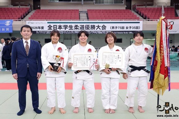El equipo femenino de judo de la Universidad de Educación de Osaka posando con judogi blanco, trofeos, diplomas y un estandarte junto a su entrenador en el Nippon Budokan.