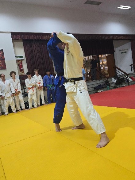 Un judoka con judogi blanco y cinturón negro eleva el brazo de su oponente con judogi azul imponiendo un agarre de Kumi-kata sobre el tatami.