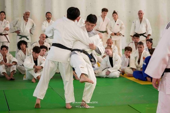 Dos judokas con judogi blanco ejecutando una técnica de desequilibrio y elevación de pierna frente a un grupo de estudiantes y profesores sentados alrededor del tatami.