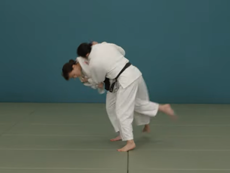 Una judoka con judogi blanco y cinturón negro elevando la pierna y traccionando la solapa para proyectar a su compañera en un estudio técnico sobre tatami.