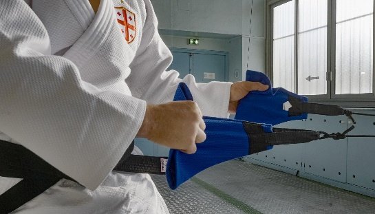 Plano cerrado de un judoka sujetando agarres de judogi azul conectados a tensores elásticos para entrenamiento de tracción.