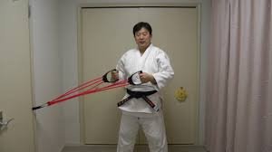 Judoka con judogi blanco y cinturón negro realizando un ejercicio de tracción frontal con tubos elásticos rojos anclados a una puerta.