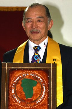 Retrato institucional del maestro de judo Tsuneo Tomita con traje formal, estola amarilla y sosteniendo la placa conmemorativa del Salón de la Fama del Deporte Riopedrense.