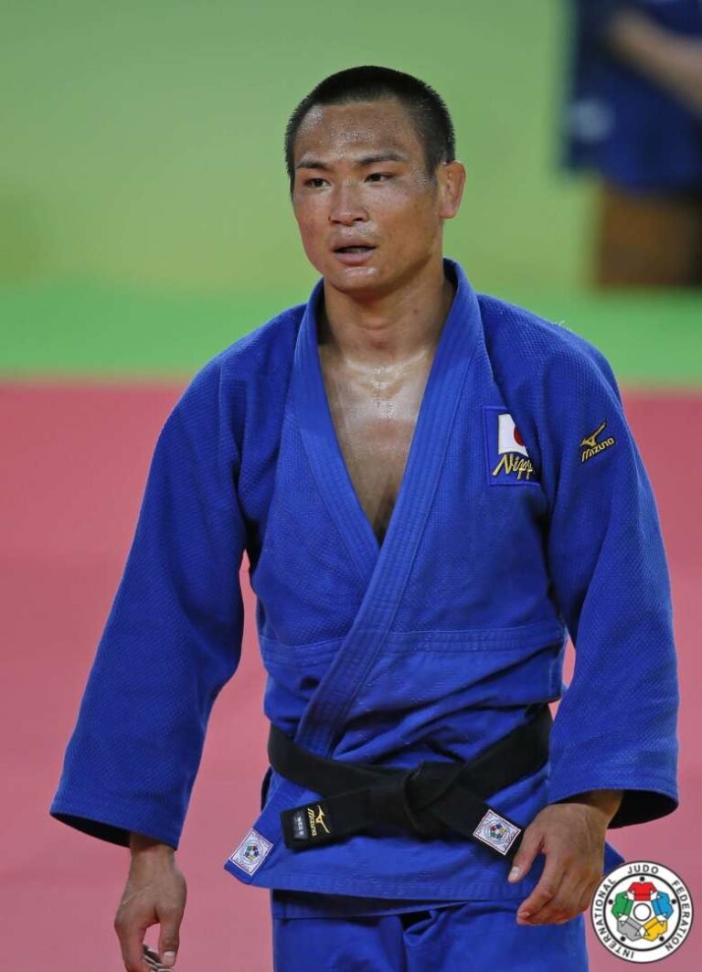 Plano medio del judoka japonés Masashi Ebinuma en el tatami luciendo judogi azul con el parche de Nippon, cinturón negro y la insignia de la IJF.