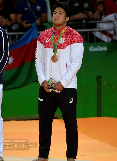 El judoka japonés Ryunosuke Haga parado sobre el podio olímpico vistiendo la indumentaria oficial de Japón y sosteniendo su medalla de bronce en Río 2016.