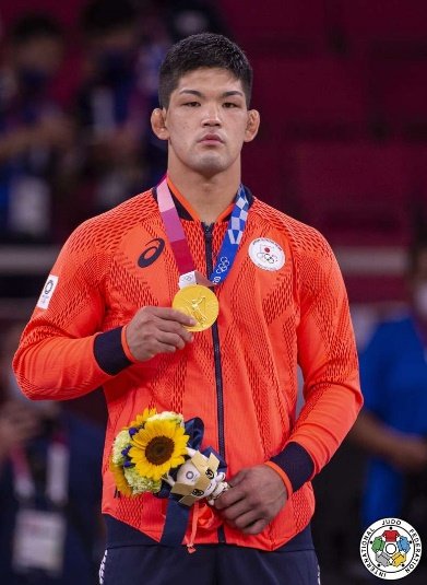 Shohei Ono en el podio de Tokio 2020 sosteniendo su medalla de oro olímpica y el ramo floral oficial vistiendo la chaqueta roja de Japón.