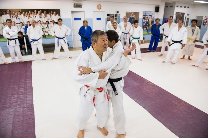 Maestro de judo con cinturón rojo y blanco demostrando una técnica de agarre y postura en el tatami ante un grupo de estudiantes.