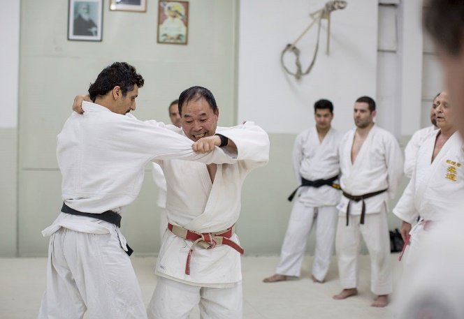 Maestro de judo con cinturón rojo y blanco enseñando una técnica de agarre Kumi-kata en el tatami ante varios judokas en el dojo.