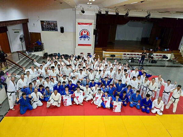 Fotografía grupal multitudinaria de judokas vistiendo judogis blancos y azules posando sonrientes con los brazos en alto sobre el tatami del Centro Okinawense.