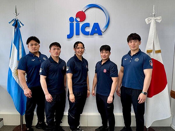 Integrantes de la delegación de judo de la Universidad de Chukyo posando alineados frente al logotipo de JICA, flanqueados por las banderas de Argentina y Japón.