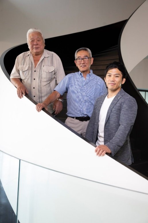 Tres maestros e instructores japoneses de judo, incluidos Kazuhiro Mikami y Hiroshi Katanishi, posando sonrientes en una escalera caracol moderna de diseño blanco y negro.